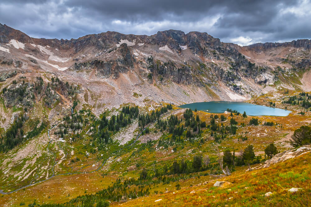 Green and brown grass, with some pine trees sparsely populating the landscape, and a tiny blue lake at the foot of mountains on a remote Grand Teton hike.