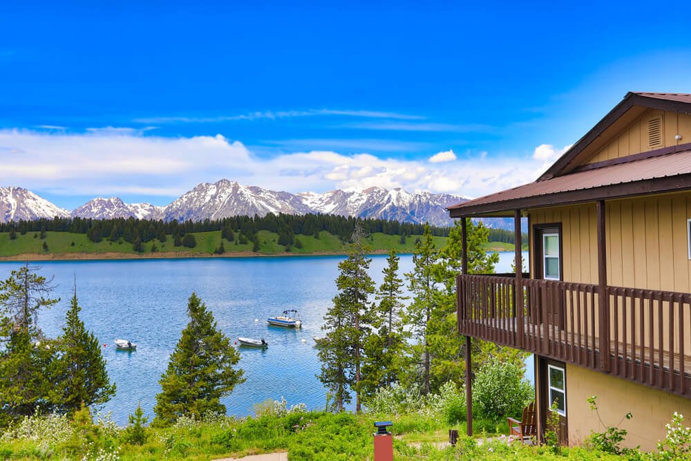 A two-story cabin overlooking a lake in Grand Teton National Park, surrounded by mountains and trees, with a few boats out on the lake on a sunny day.