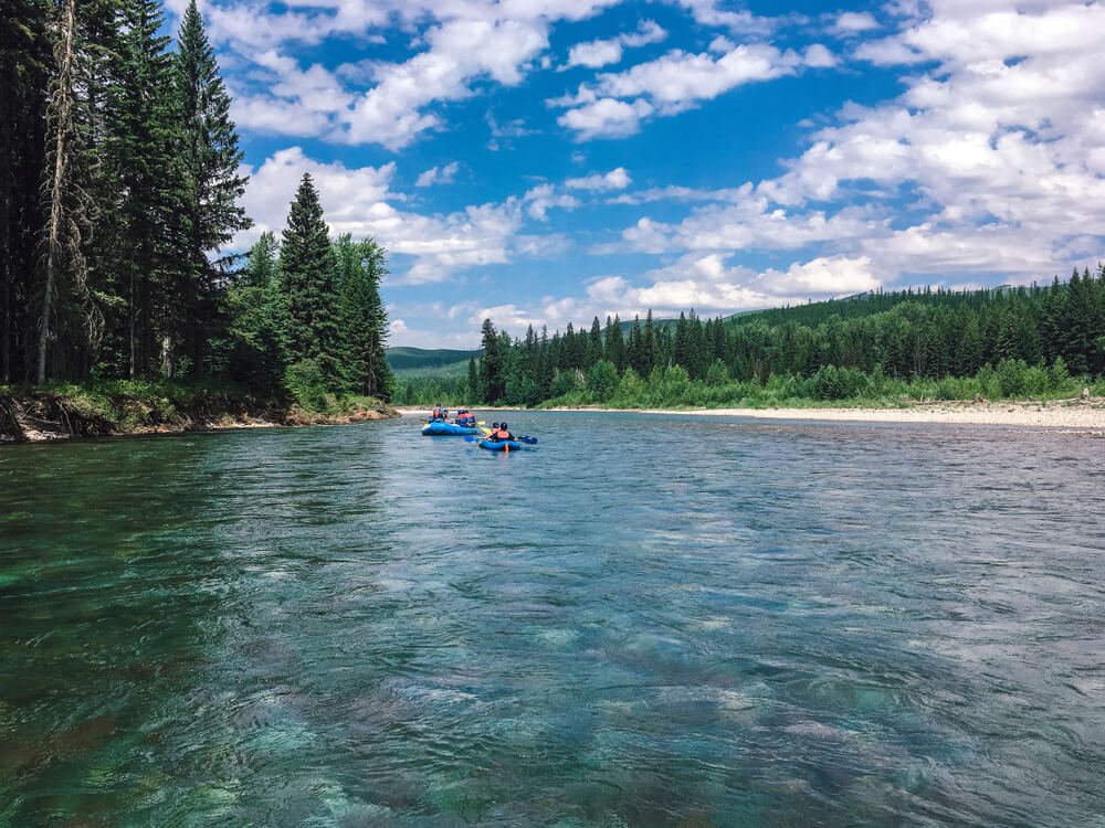 Two rafts ahead on the Flathead River, which is calm, turquoise and surrounded by trees and hills on a sunny day in Glacier National park