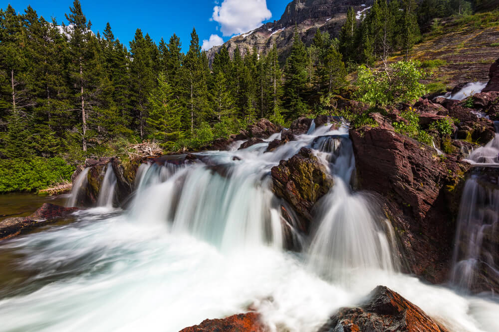 Small cascade over red rocks at Redrock Falls, a popular hike in Glacier National Park, surrounded by trees and blue sky.