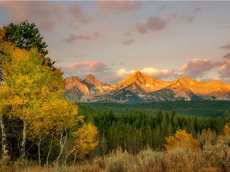 Autumn is the best time for an Idaho road trip: yellow trees, green evergreens, and orange mountain tops on a partly cloudy day.