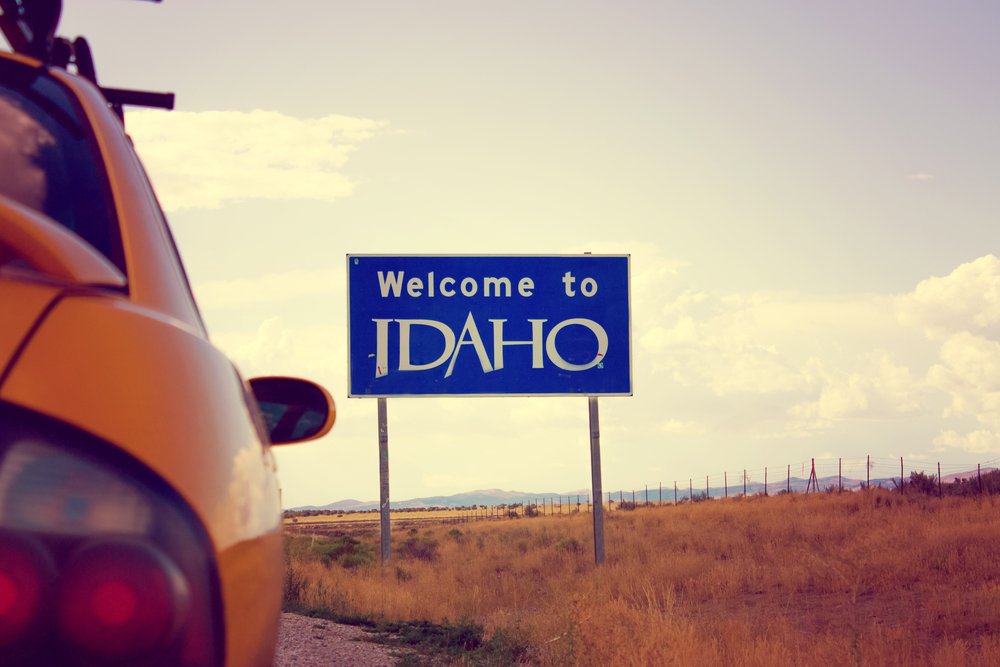 Sepia-toned photo which shows the back of a car approaching a sign which says "welcome to Idaho" on an Idaho road trip with a field in the background.