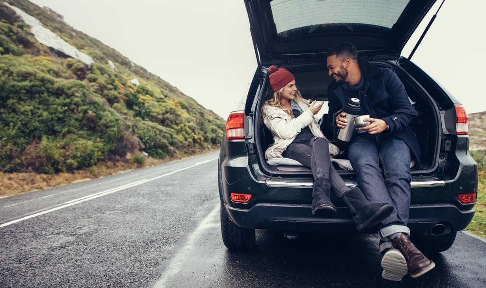 Interracial couple (white woman and Black man) sharing a thermos of coffee while pulled over on the road while wearing cold-weather clothing, sitting in the back of their car.