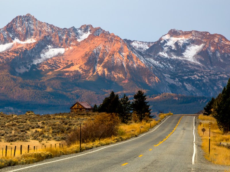 An open road with yellow grass on the sides of it, a barn on one side of the road, and mountains with some snow lit up orange by the sun.