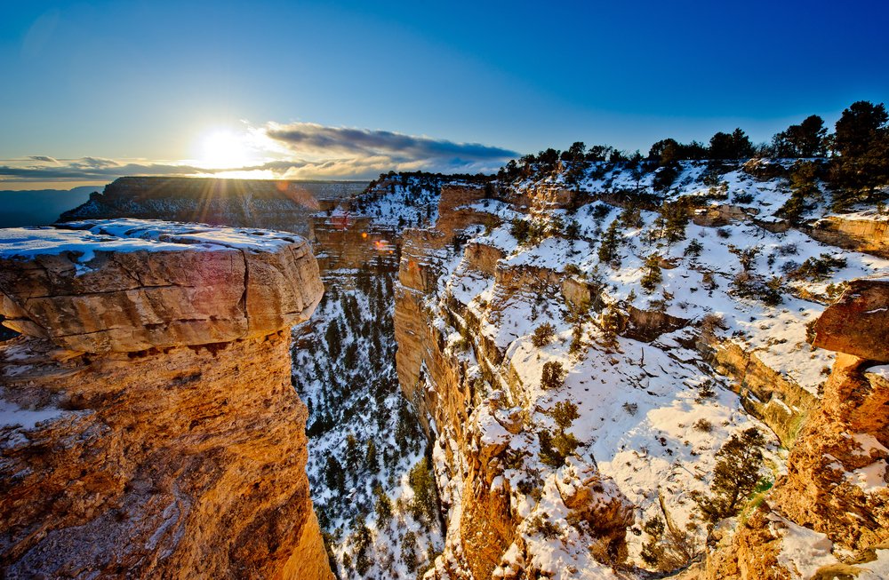 Snow covered landscape at the Grand Canyon in winter, red rocks with patches of white snow with the sun rising above the canyon at sunrise.