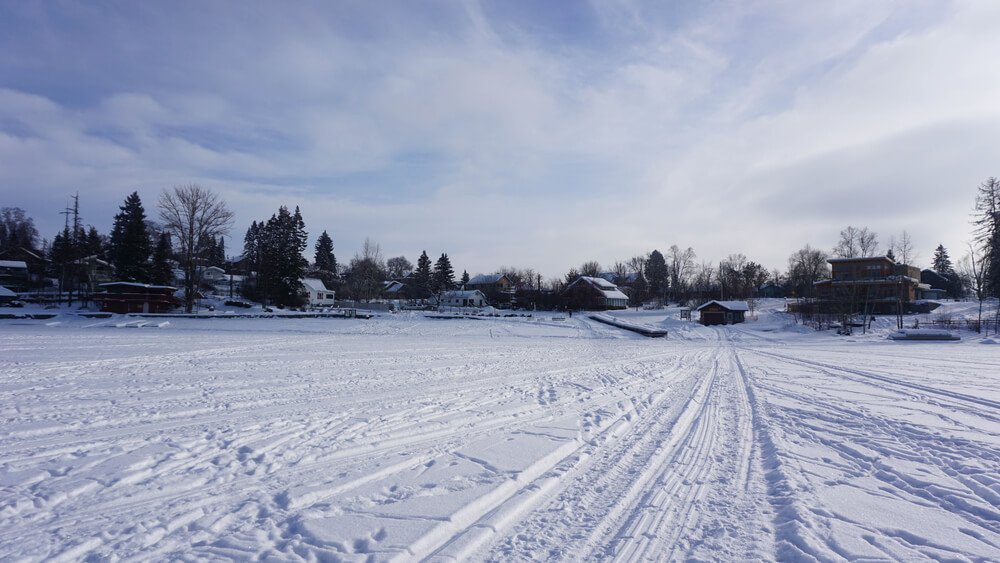 Snowmobile tracks on the frozen lake at Whitefish Lake in Whitefish, MT with houses and lodges in the background.