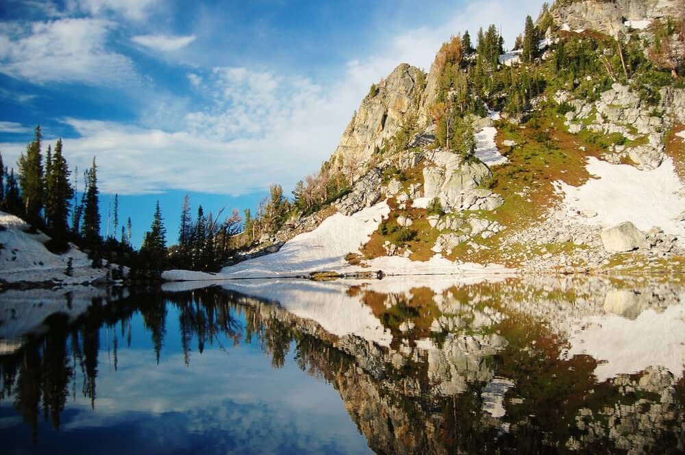 Still lake reflecting back the mountains and foliage above it, covered in some light snow that hasn't yet melted.