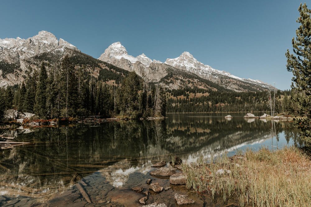 Still lake at Taggart Lake reflecting the Teton range above it, surrounded by trees, rocks, and foliage.