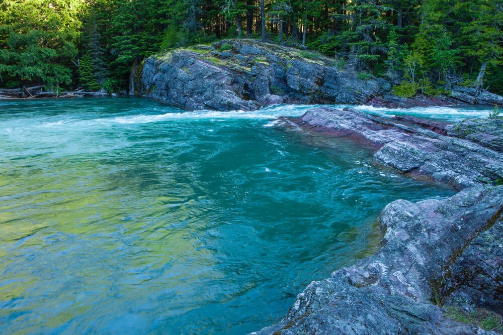 Brilliant emerald-turquoise waters at Upper McDonald Creek, surrounded by rocks covered in green moss and trees.