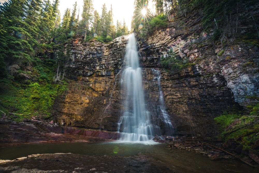 Waterfall cascading over a cliff, with trees surrounded the edges of the cliff with a small sunburst poking through the trees.