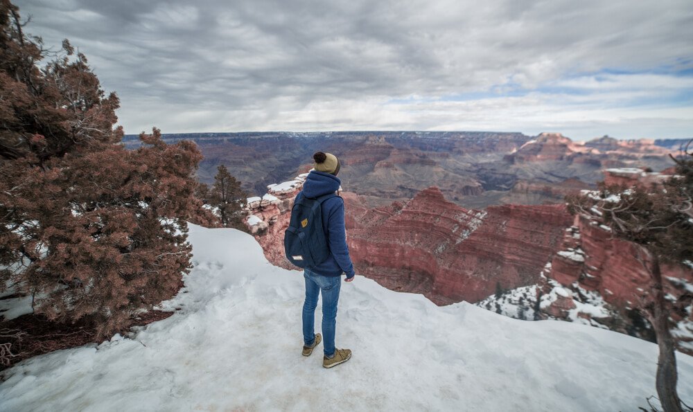 Man standing a few feet from the edge of the Grand Canyon in the snow, wearing a hat, sweater, and jeans.