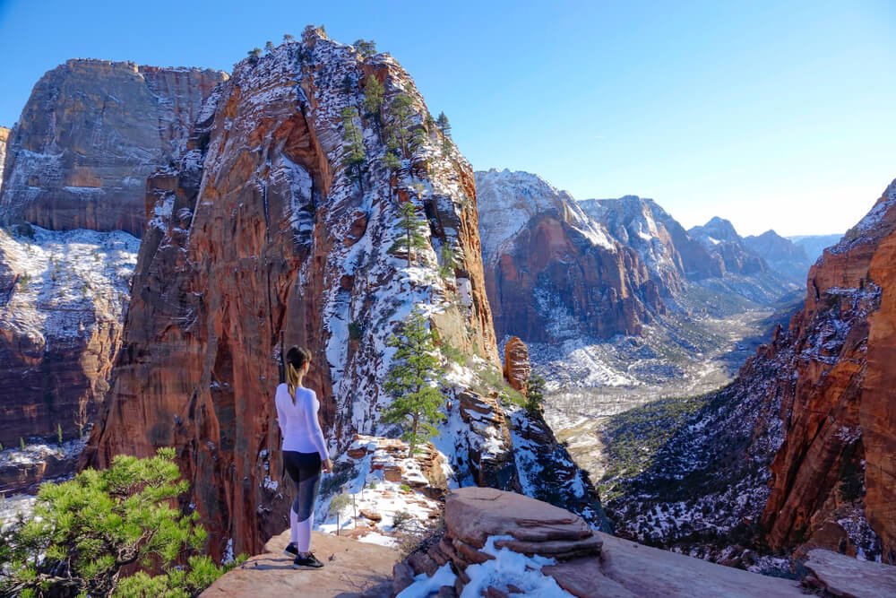 A woman in a lavender top and leggings hiking the ridge of Angel's Landing covered in a light snow, looking over a snow-covered valley in Zion in winter.