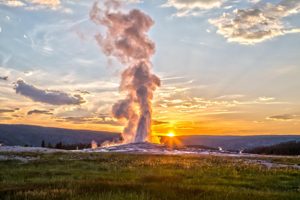 The Old Faithful geyser at sunset, a plume of steam shooting high into the air as the sun sets behind it, a classic sight on any Yellowstone itinerary.