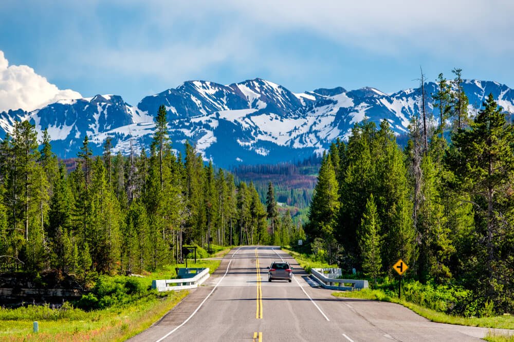 A car on the road heading towards snow-covered mountains on a Yellowstone road trip between Yellowstone and Grand Teton National Parks.