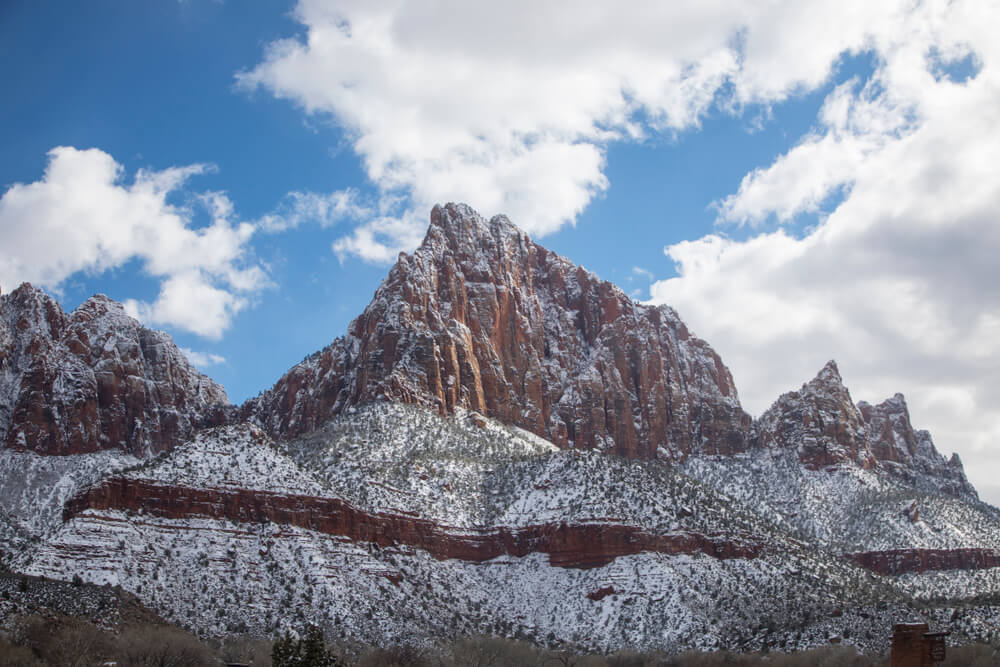 Snow-covered mountain near the Watchman Campground, with a partly cloudy sky in the background. Red rock showing behind the snow. 