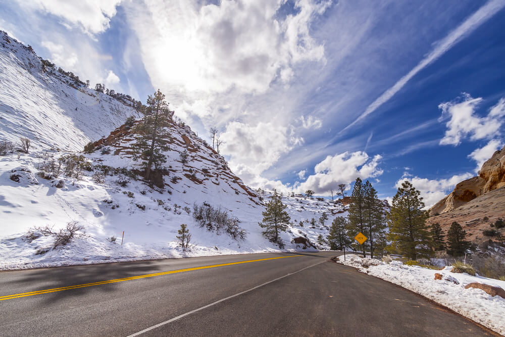 Snow-covered canyon walls with a sunny sky with some clouds, a plowed road that is empty winding through Zion in winter.