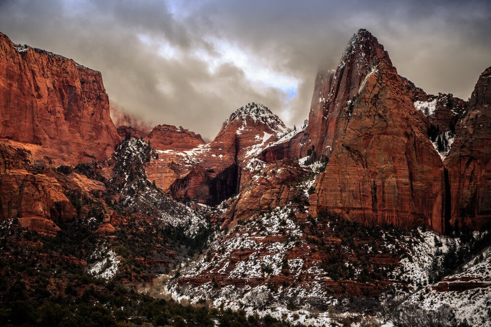 Kolob Canyon walls covered in a light snow which shows from underneath the red rock, a stormy sky with dark clouds above.