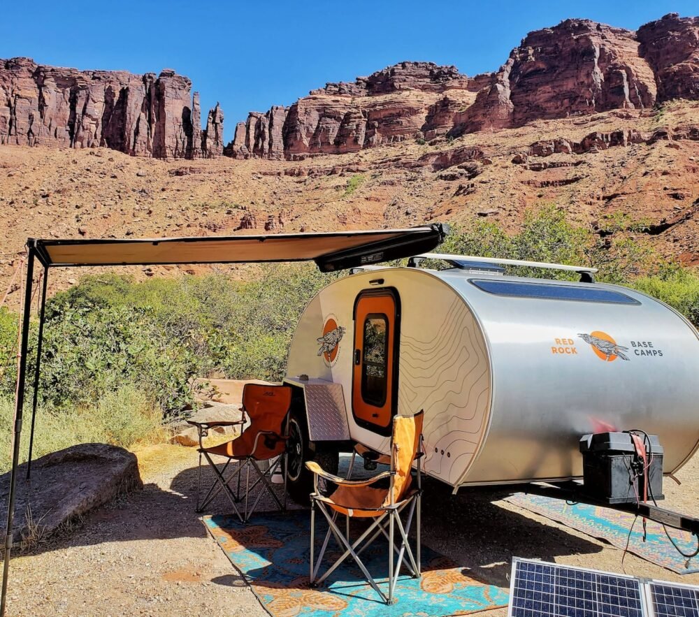 A silver teardrop trailer with an awning over it, with two orange camping chairs, next to a solar panel in a beautiful red rock landscape typical of Moab.