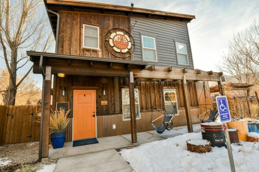 Wooden two-story house with orange door and two porch swings in front with a sign on the Moab airbnb which reads "Moab Digs"