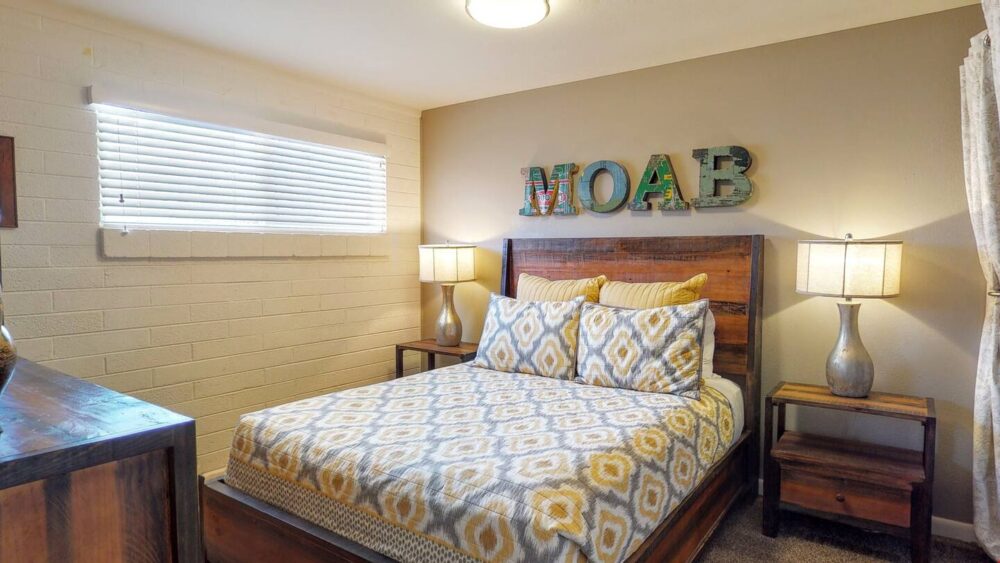 Bed with colorful yellow and gray bedspread with the words "MOAB" in block letters written above it with an exposed brick wall painted white to one side.
