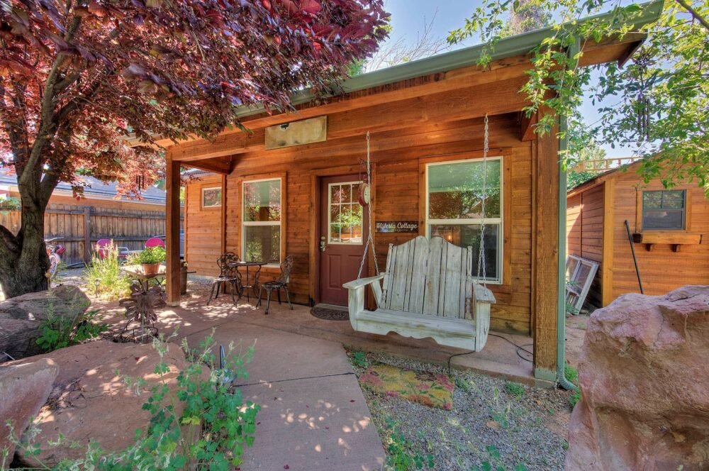 A romantic, whimsical cottage surrounded by a tree and flowers, with a pastel green porch swing and a small dining table in front of the cottage in Moab.