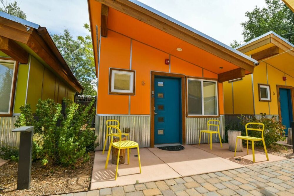 A brilliant coral pink orange house with four yellow chairs in front and a slanted tin roof, next to other colorful houses (green and yellow).