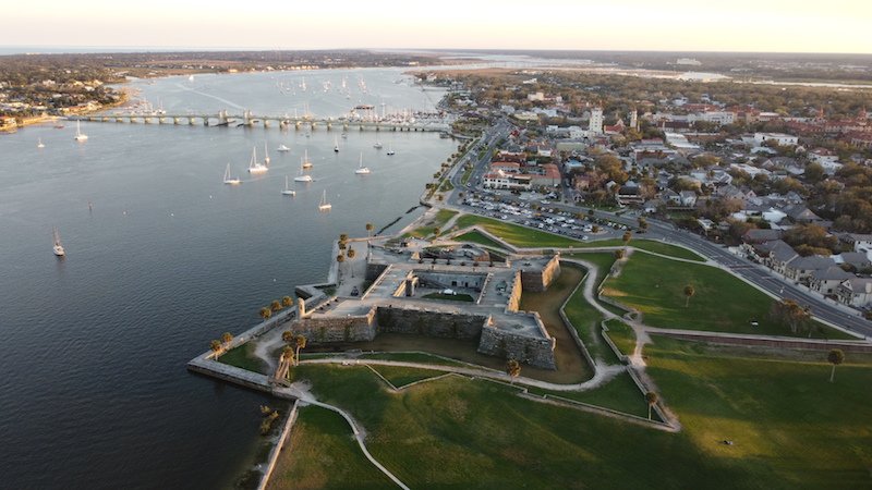 A drone photo of of a fort shown in the shape of a star, with the view of water and a bridge and sailboats in St Augustine FL