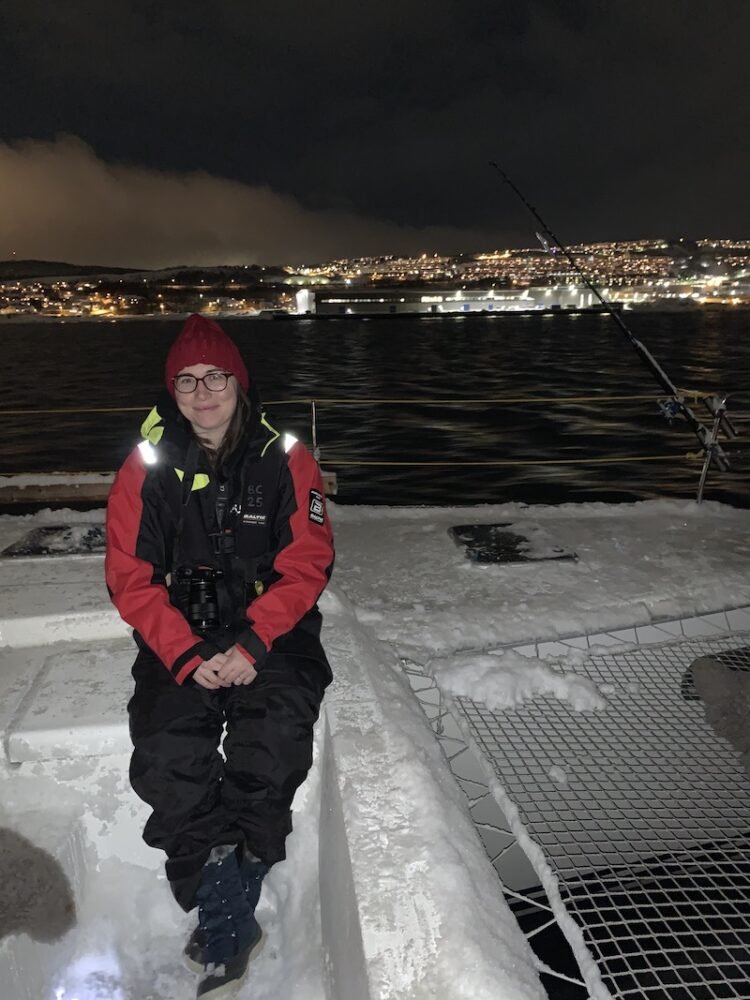 Allison Green wearing a red thermal suit while sitting on a snow-covered catamaran sailing in the Norwegian fjords, hoping to see the Northern lights. There are the lights of the city of Tromso visible in the background and snow on the catamaran, showing that it is deep winter!