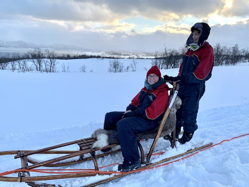 Allison Green sitting on a dog sled with a fellow solo traveler, wearing a red hat and blue scarf and a red and blue expedition suit