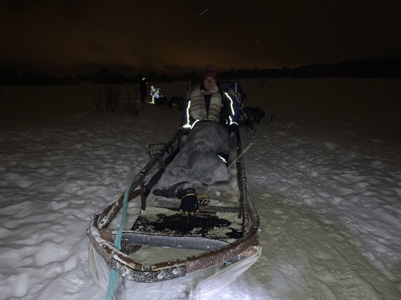 Allison Green in the dark wearing a reflective snow suit with a scarf while on a Northern lights and husky tour