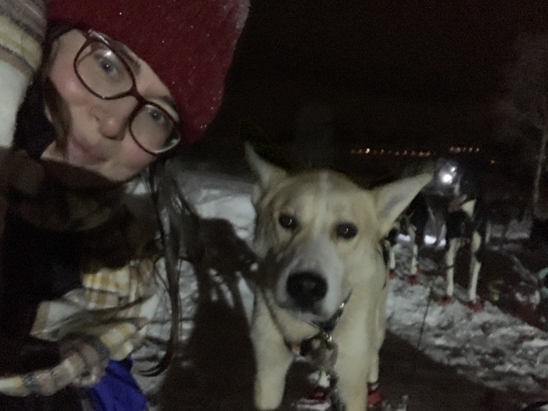 Allison Green wearing a hat, scarf, and glasses, next to a dog on the dog sledding team. The night sky is very dark and the photo is a little blurry.