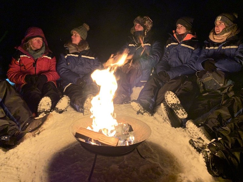 People in thermal suits sitting around the fire while waiting for the aurora to appear on a Northern lights tour in Tromso.