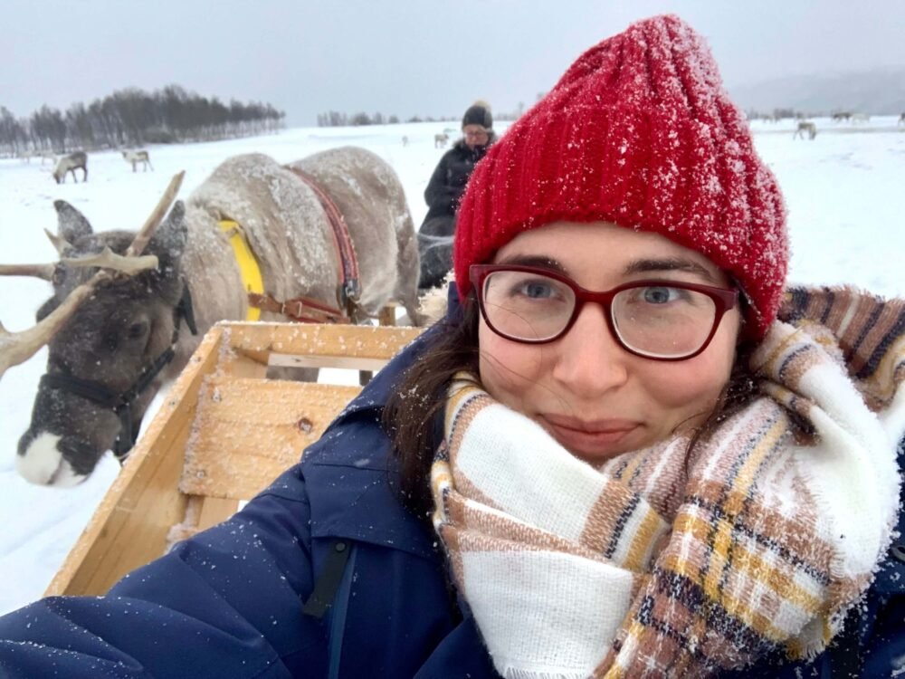 Allison Green wearing glasses, a scarf, a winter jacket and a red hat while sitting on a sled in Tromso doing a reindeer sledding experience