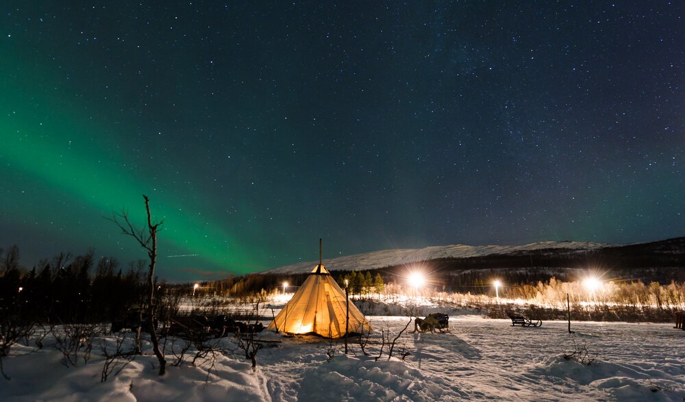 A Sami reindeer camp with an aurora over it and sleighs visible in distance