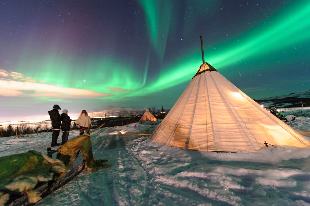 A sami reindeer camp with the aurora over it and people looking at the aurora and a lit-up lavvu or Sami tent