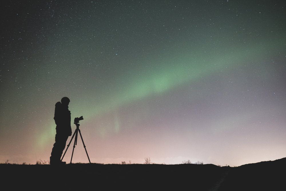 a man photographing the northern lights with a camera and a tripod with the aurora visible behind him