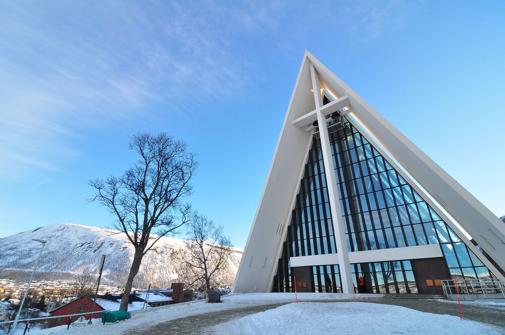 Large white church with a big cross and snow and views of mountains in distance on a sunny winter day. Arctic Cathedral is a must on a Tromso itinerary