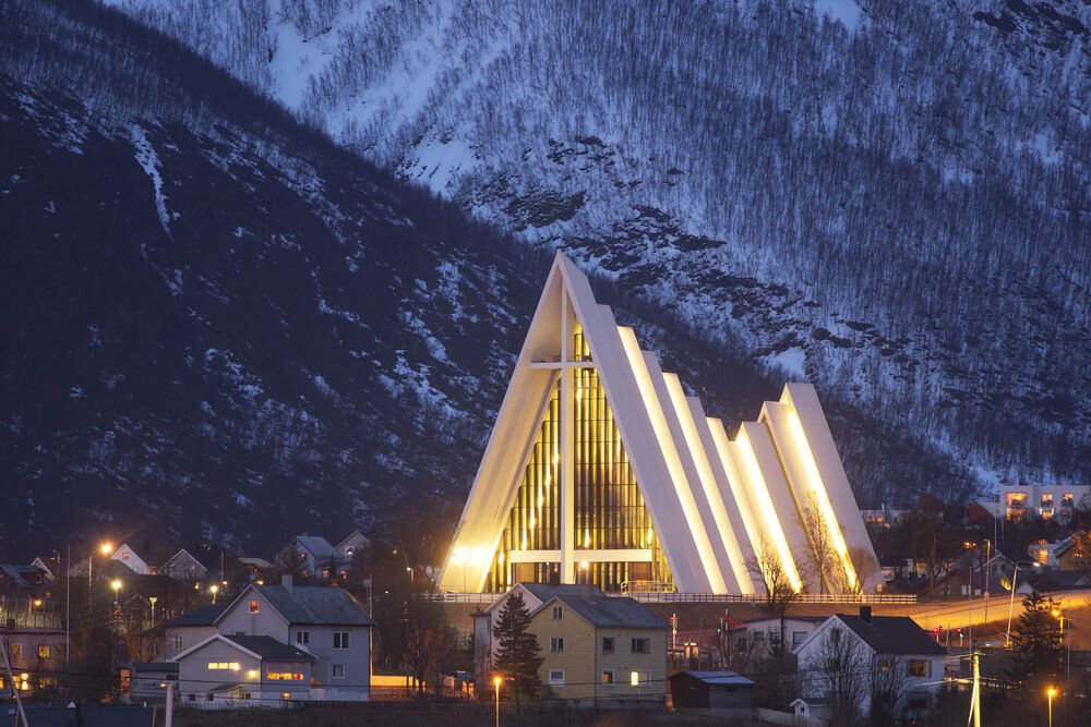 The lit-up Triangular architecture of the Tromso Arctic Cathedral against a mountain backdrop in the snow in winter.
