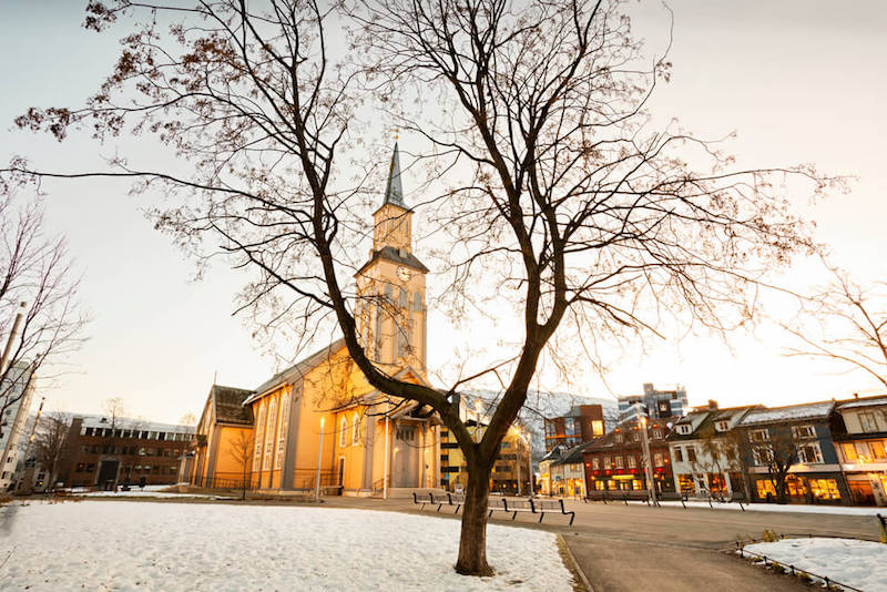 Brownish-tan wooden cathedral in a square in Tromso Norway in winter with snow on the ground and buildings lit up in evening