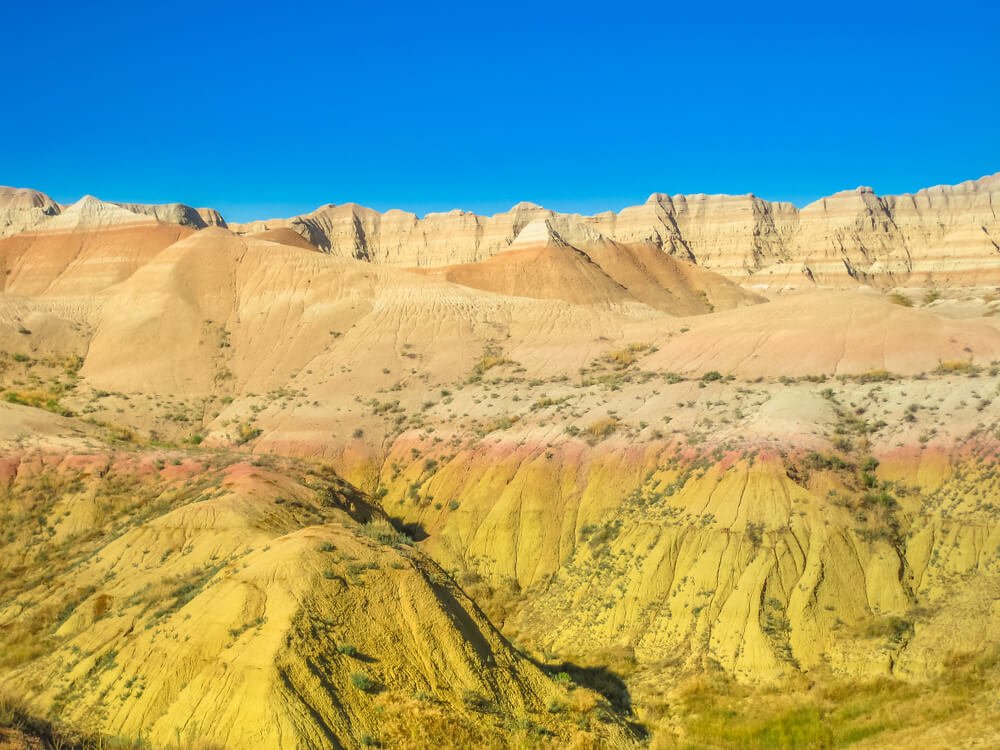Yellow mounds in Badlands National Park with stripes of orange, tan, and pinkish-red with a clear blue sky behind it