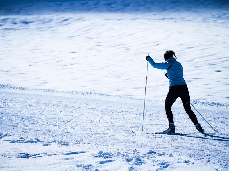 woman in a blue shirt and snow pants with cross country skis exploring in the snow