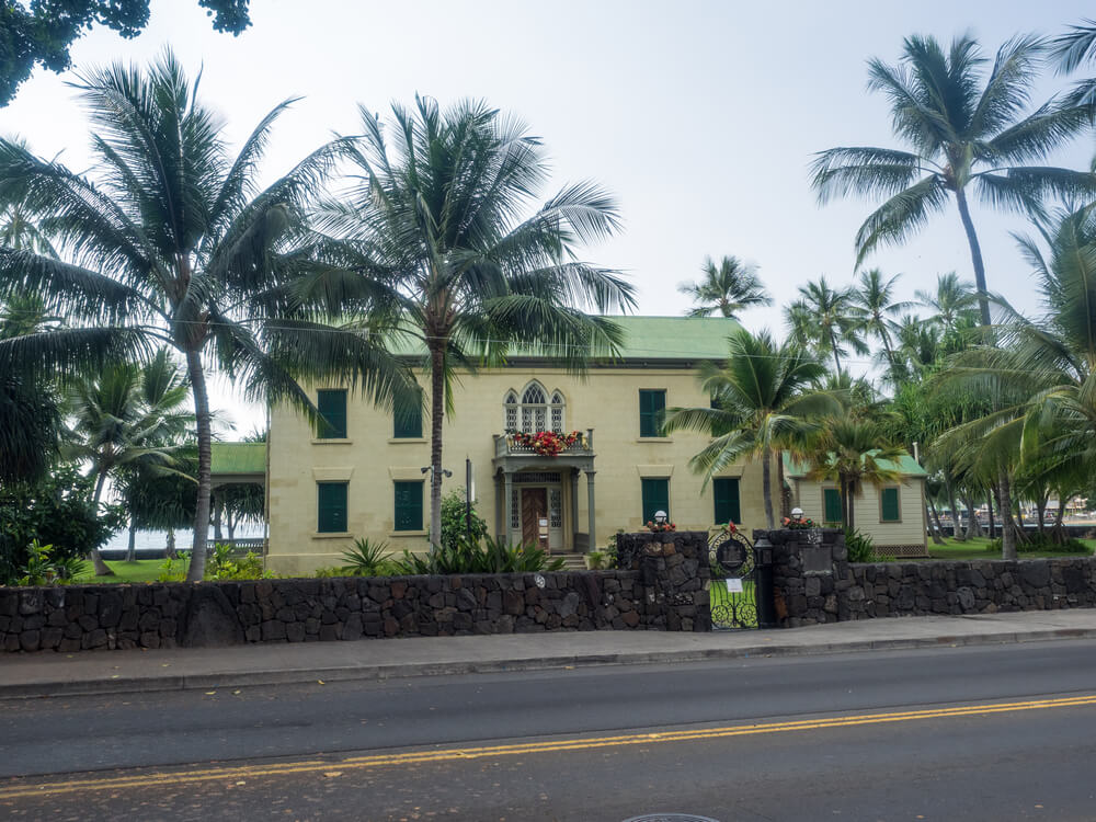 yellow toned historic building of hulihee palace on the road with palm trees in front of the ocean