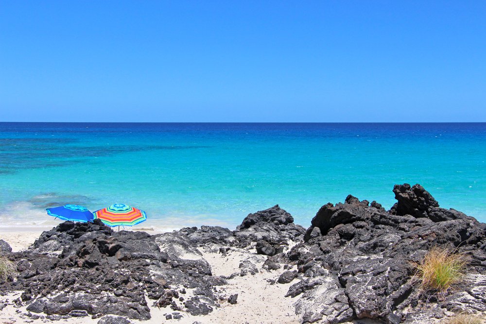 two colorful beach umbrellas behind black lava rock with beautiful turquoise sea and white sand in the background