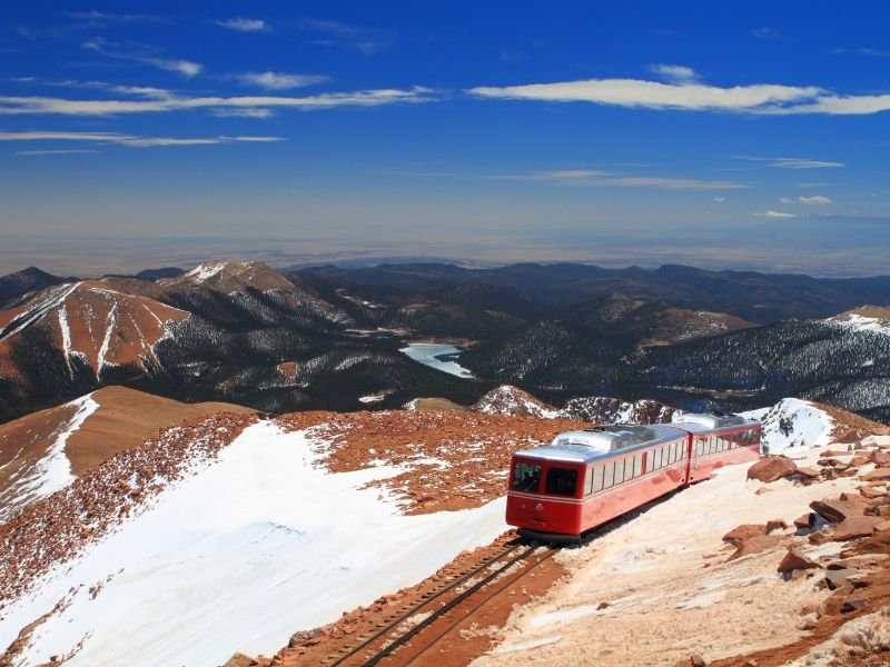 famous red railway car with a silver roof ascending pikes peak with view of mountains with light dusting of snow in the distant background