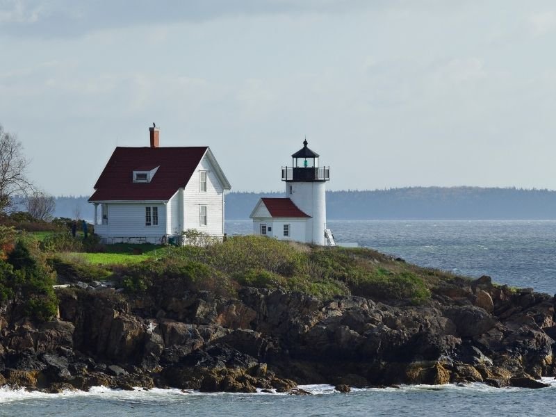 a beautiful white lighthouse with a red roof and lightkeeper house on a rocky outcropping in the bay near camden