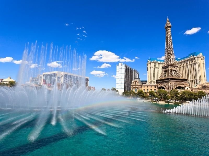 fountain in front of the bellagio during the day time making a small rainbow prism in the water with the eiffel tower visible in the background