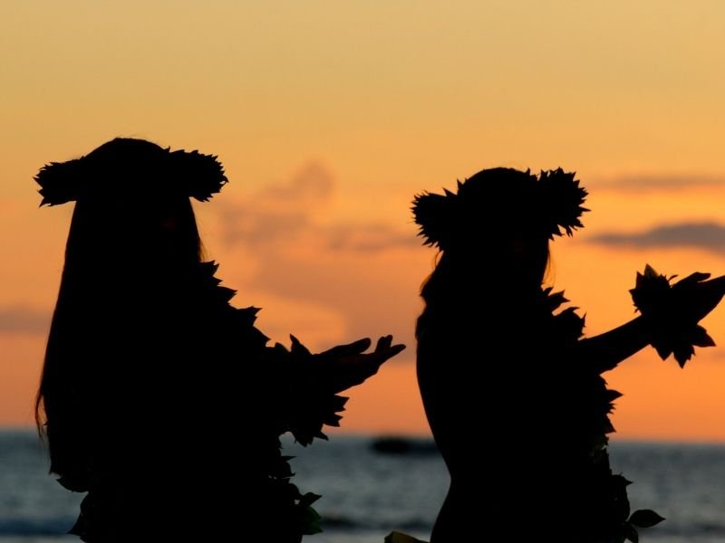 two female figures silhouetted against the orange sky at sunset doing a hula dance at a luau on this big island itinerary