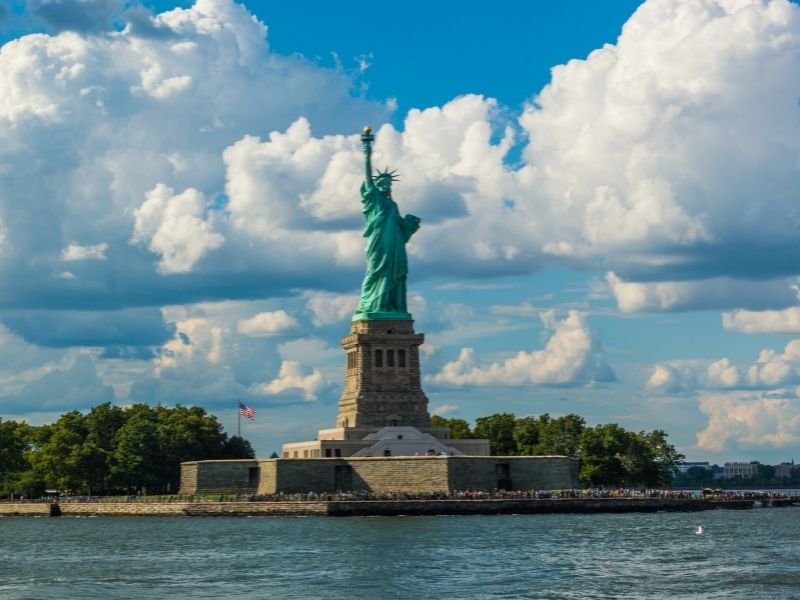 faraway view of statue of liberty from on the ferry with lots of people crowding around in the distance