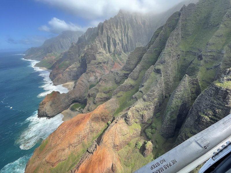 View of the Na Pali Coast as seen from a doors off helicopter. Red rock and green landscape and rugged coastline and ocean water.