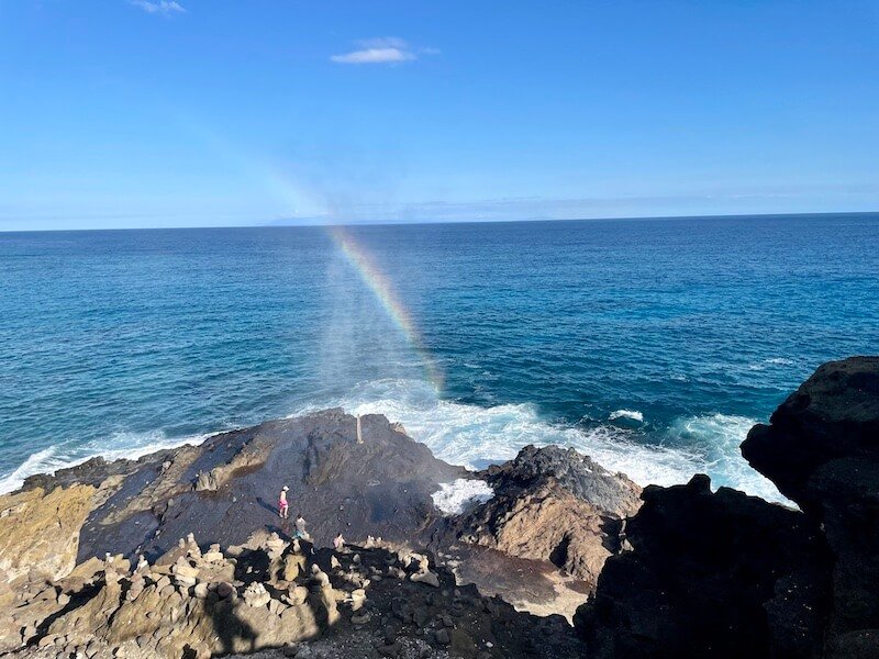 water from a natural 'blowhole' coming out of a lava tube and making a rainbow in the sun